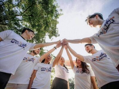 Group of people in matching white T-shirts stacking hands together in a team huddle outdoors